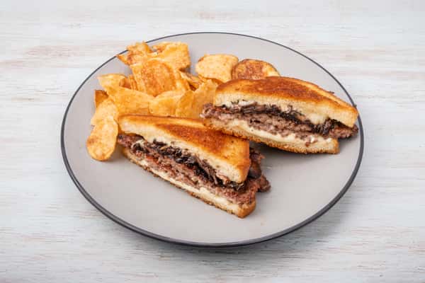 Truffle mushroom patty melt sandwich with melted cheese on grilled bread, cut in half and served with potato chips on a gray plate at a restaurant