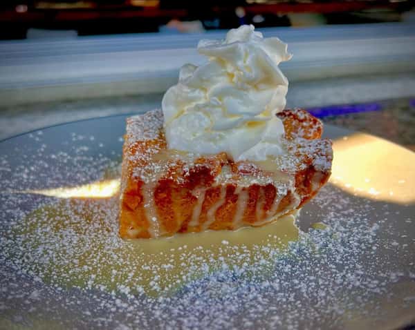 Warm bread pudding topped with whipped cream, powdered sugar, and sweet glaze on a dessert plate