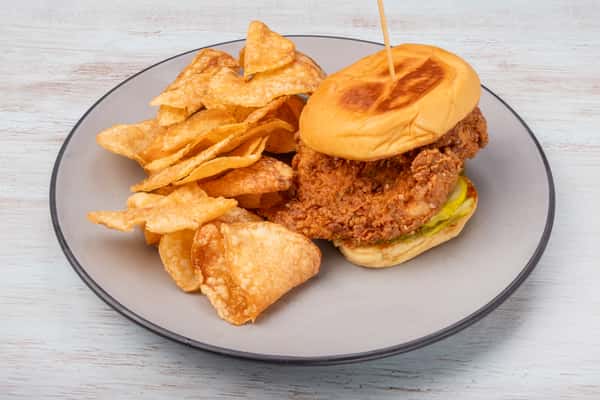 Fried chicken sandwich with pickles on a toasted bun, secured with a skewer pick, served with potato chips on a gray plate at a restaurant