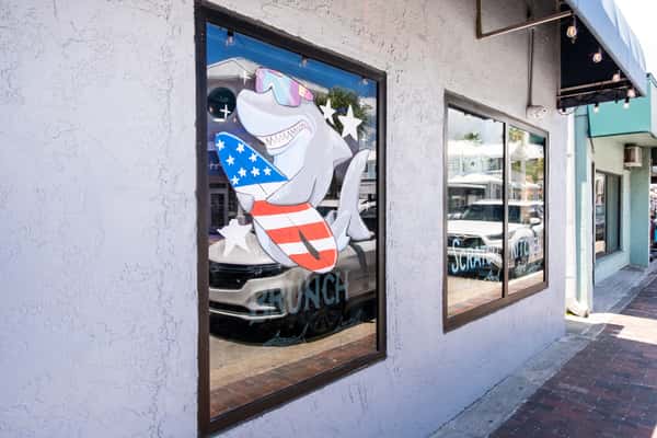 Storefront window mural of a shark mascot in patriotic American flag attire, painted on a restaurant's exterior window