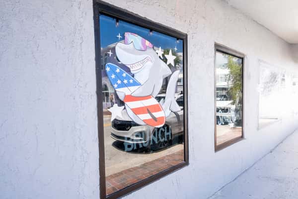 Window mural of a shark mascot in patriotic attire painted on the exterior of a restaurant, with brunch signage