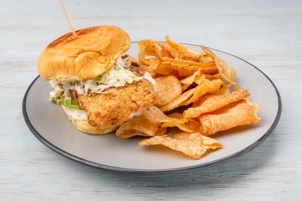 Crispy fried fish sandwich topped with coleslaw on a bun, secured with a skewer pick, served with potato chips on a gray plate at a restaurant