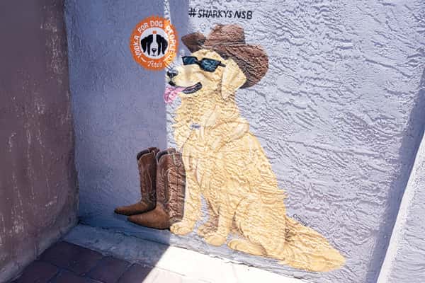 Mural painting of a dog wearing sunglasses and a hat next to a pair of cowboy boots on an exterior wall at a restaurant