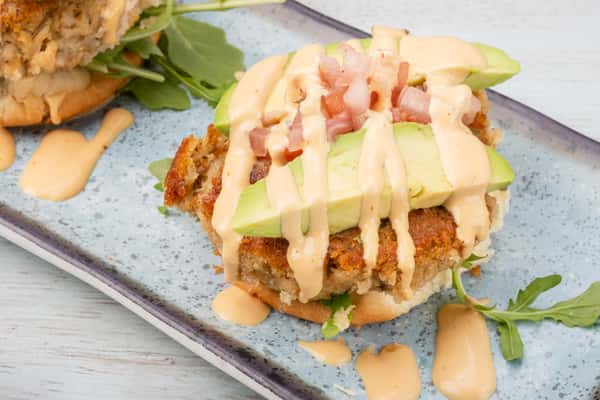 Crab cake sliders topped with sliced avocado, diced tomato, and a drizzle of sauce, garnished with greens on a rectangular slate plate at a restaurant
