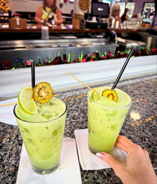 Two bright green kiwi lime cocktails served at a restaurant bar with fresh lime wedges and dried kiwi garnish