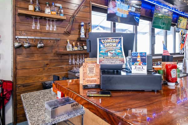 Host stand with a cash register, wine glasses hanging above, and a comedy show flyer on a wooden bar counter inside a restaurant