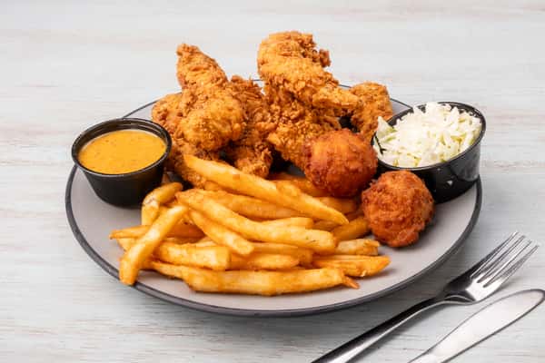 Crispy fried chicken tenders served with french fries, coleslaw, a fried hush puppy, and dipping sauces on a gray plate at a restaurant