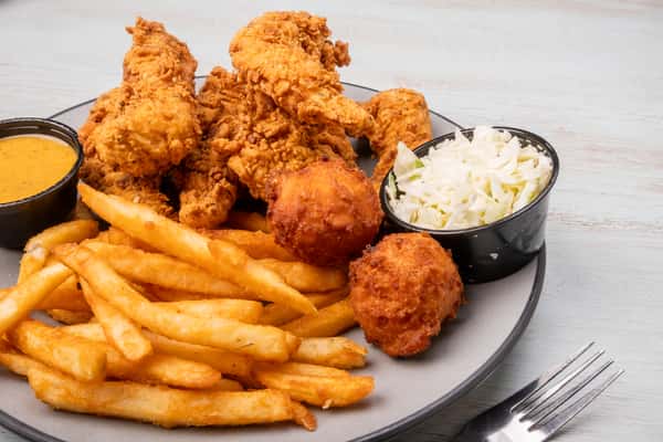 Crispy fried chicken tenders served with french fries, coleslaw, and a fried hush puppy on a gray plate at a restaurant