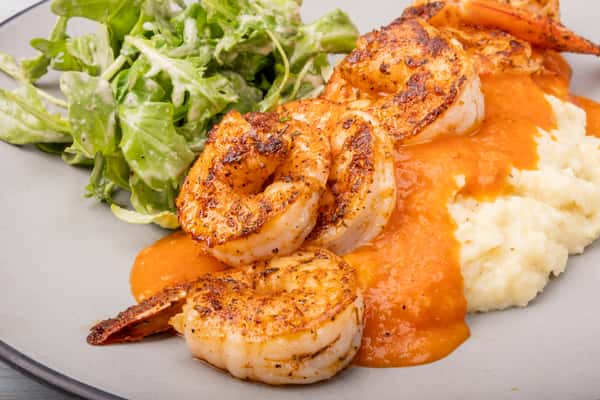 Grilled shrimp over creamy grits topped with a spicy sauce, served with a side salad on a gray plate at a restaurant
