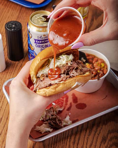 A patron adds sauce to a barbecue sandwich, piled high with pork and coleslaw. The image evokes a sense of down-home southern cooking. Text: "Austin Eastciders Original Dry Cider."