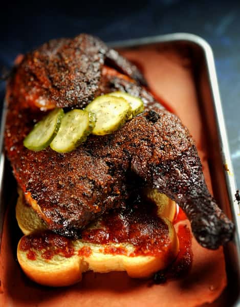 Hot chicken on a bun, served on butcher paper in a metal tray, is topped with dill pickle slices. The rich lighting shows off the depth of color in the bird's seasoning.