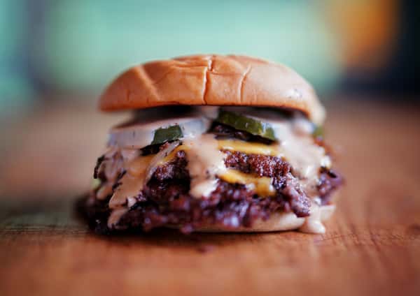 A delectable burger is centered on a wooden table, topped with sauce and pickles, and photographed with a shallow depth of field. Perfect for showcasing the restaurant's enticing gallery.