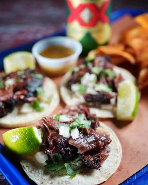 Three beef tacos sit on a blue tray, garnished with cilantro, onion, and lime. A small container of sauce and potato chips rest in the background.