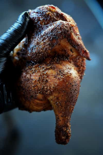 A whole smoked chicken with crispy skin is held by a person wearing a black glove. The chicken is golden brown and heavily seasoned, ready to be enjoyed.
