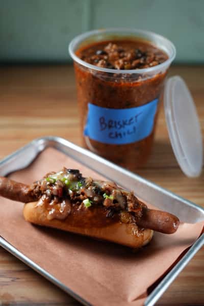 A brisket chili dog and container of brisket chili rest on a metal tray. The container is labeled "Brisket Chili".