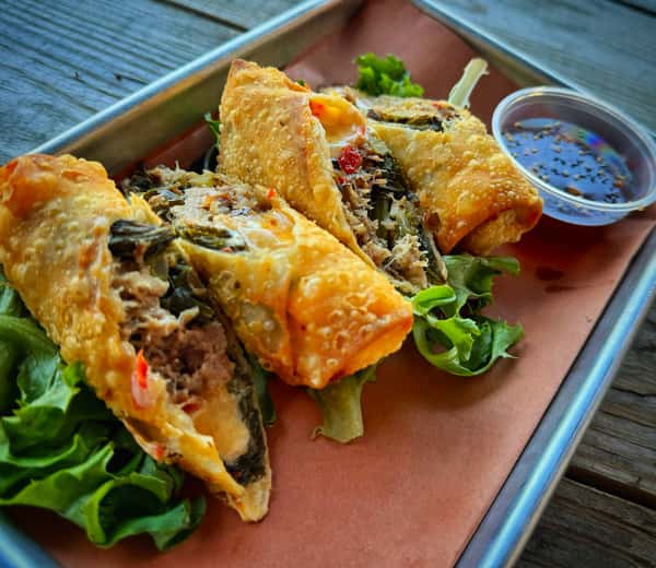Crispy egg rolls on lettuce are served with a small dish of dipping sauce. The egg rolls appear to be sliced open to show the savory filling.
