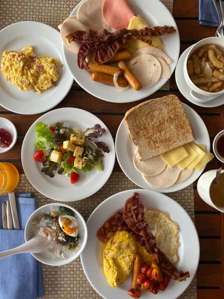 Overhead view of a large breakfast spread with eggs, bacon, sausages, sliced meats and cheese, toast, salad, rice porridge, soup, and drinks.
