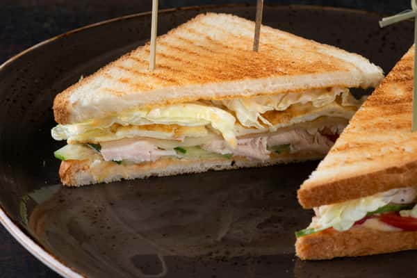 Grilled sandwich made with toasted white bread, layered with slices of chicken, cucumber, lettuce, tomato, and egg, held together with skewers.