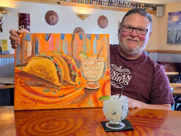 Man seated at a table holding a colorful painting, with restaurant decor behind him.