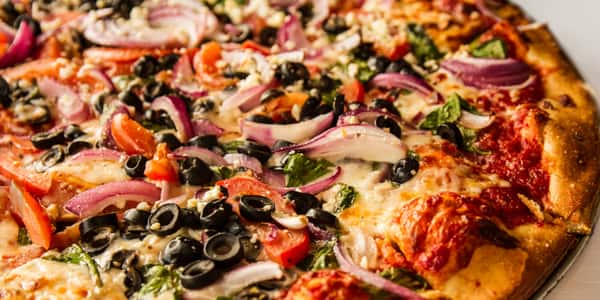 Close-up of freshly baked vegetable pizza topped with black olives, red onions, tomatoes, spinach, and melted mozzarella on a golden crust.