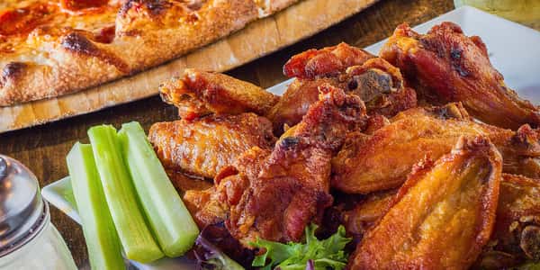 Plate of crispy buffalo-style chicken wings served with fresh celery sticks and dipping sauce, with a wood-fired pizza in the background at a casual restaurant bar.