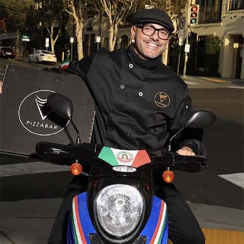Chef in black uniform riding a scooter with a PizzaBar delivery box, smiling while making a pizza delivery in an urban street setting.