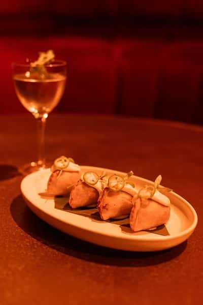 Elegant appetizer trio served on a modern ceramic plate, featuring bite-sized tacos or taquitos wrapped in golden tortillas and topped with thin jalapeño slices and herbs, paired with a clear cocktail in a coupe glass—set against a dimly lit, upscale dining ambiance.