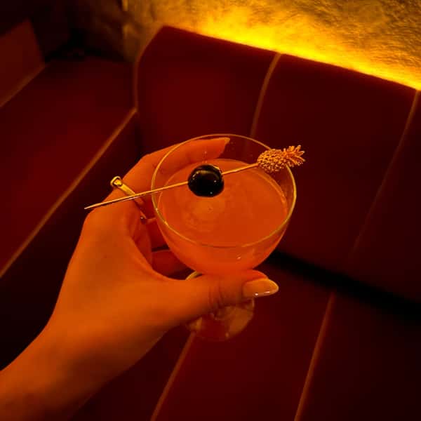 Close-up of a hand holding an elegant orange-hued cocktail garnished with a dark cherry and golden pineapple pick, against a velvet booth and moody amber lighting—ideal for upscale bar or lounge promotions.