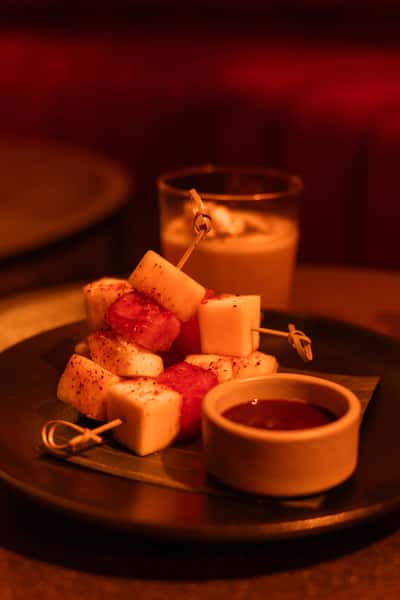 Close-up of skewered cheese and watermelon cubes sprinkled with spices, served with a dark dipping sauce on a ceramic plate—ideal for bar snacks or modern small plate menus.