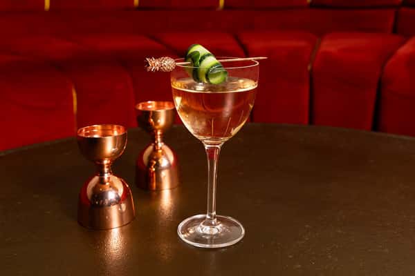 Sophisticated clear cocktail garnished with ribboned cucumber and a golden pineapple pick, set on a dark table alongside copper jiggers, with plush red lounge seating in the background—evoking a refined cocktail lounge setting.