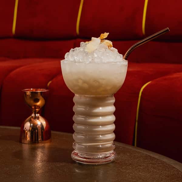 Frosty tropical cocktail served in a sculpted glass, topped with shaved coconut and crushed ice, with a metal straw on a dark table.