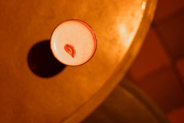 Top-down view of a creamy craft cocktail with foam art resembling lips, served on a golden-toned table under warm, ambient lighting—perfect for a stylish speakeasy or upscale bar atmosphere.