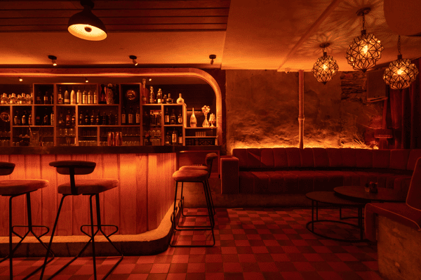 Moody cocktail lounge with a glowing backlit bar, high velvet bar stools, and geometric pendant lights casting warm amber tones over red-tiled floors and plush booth seating—perfect for a romantic night out or upscale evening drinks.