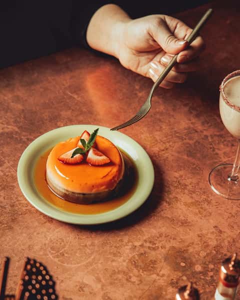 Decadent flan topped with fresh strawberries and mint, served on a ceramic plate with caramel sauce—captured on a copper-toned table for a rich, inviting dessert presentation in a Latin-inspired or upscale restaurant.