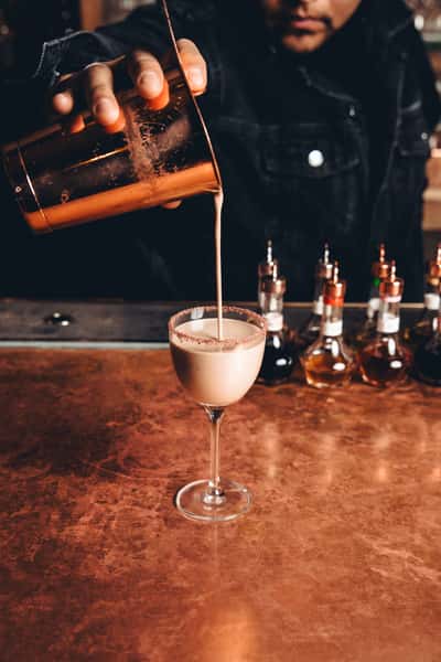 Bartender pouring a creamy cocktail from a copper shaker into a coupe glass rimmed with spice, set on a polished copper bar—evoking artisan mixology and premium bar service.