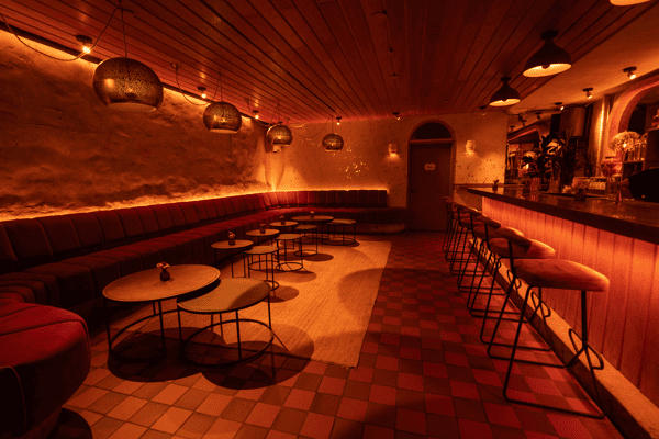 Lounge-style bar seating with plush red banquettes, small round tables, and warm pendant lighting, creating a cozy, intimate vibe for evening cocktails or small group gatherings.