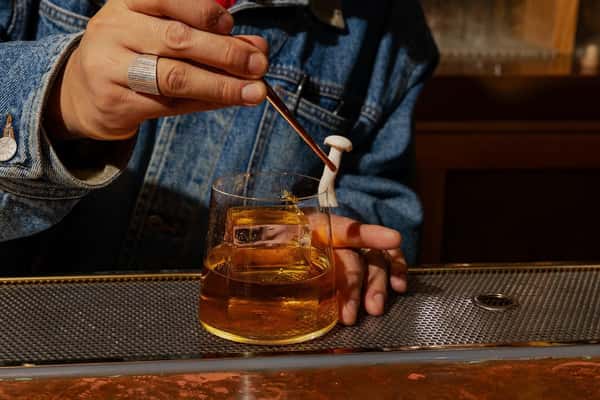 Bartender garnishing a golden-hued cocktail in a lowball glass with large ice cubes, wearing a denim jacket at a stylish bar counter.