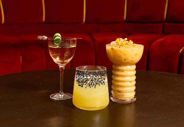 Assorted trio of craft cocktails including a cucumber-garnished martini, a yellow citrus drink with black sesame rim, and a creamy tropical cocktail in a sculpted glass—styled against red velvet seating for a modern mixology showcase.