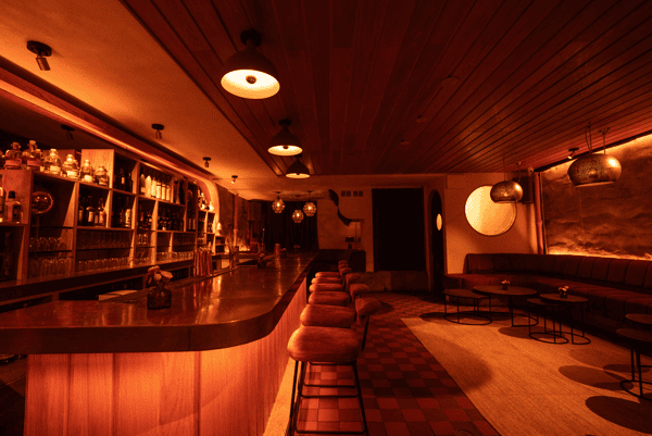 Sleek cocktail bar with a long polished wood counter, plush velvet stools, and ambient pendant lighting, set beside a cozy lounge with low tables and textured walls.
