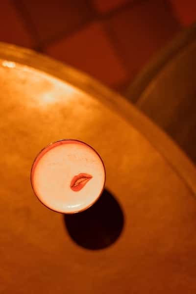 Artfully crafted cocktail with a creamy foam top, garnished with a lip-shaped edible accent, viewed from above on a moody, warmly lit bar table—highlighting a trendy mixology presentation.