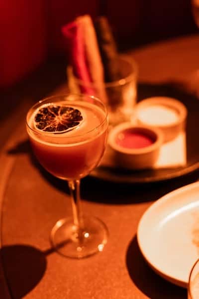 Chic cocktail in a coupe glass topped with a dried citrus wheel, accompanied by a tray with dipping sauces and colorful vegetable crudités in the background, evoking a vibrant yet refined dining experience.