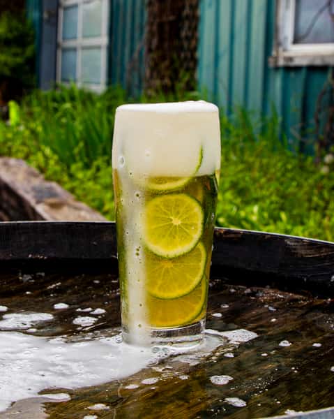 Tall glass of light beer with fresh lime slices and foamy head sitting on a wet outdoor barrel table near the brewery