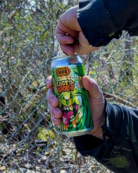 Person cracking open a can of Voodoo Brewing Liquid Hooker craft beer with foam spraying outdoors beside a chain link fence