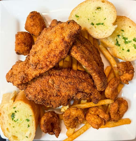 Fried fish fillets, shrimp, french fries, two hush puppies, and garlic bread on a white plate.