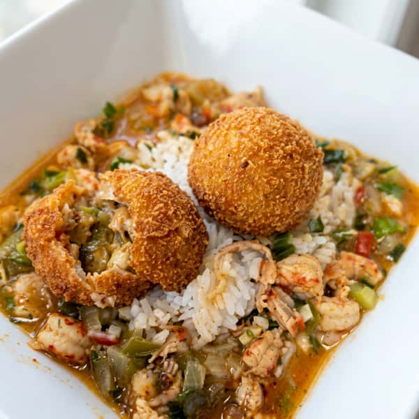 Crawfish étouffée with two golden-brown fried balls, one cracked open to show a white filling, served over rice.