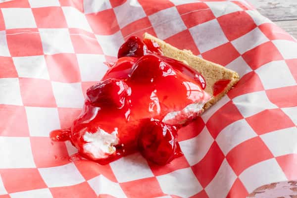 A slice of strawberry cream pie topped with glossy red strawberry sauce, served on red and white checkered paper