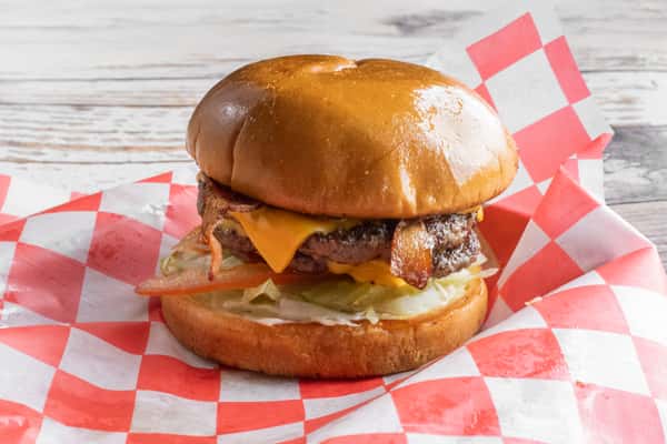 A large bacon cheeseburger with melted cheese and toppings on a toasted brioche bun, served on red and white checkered paper