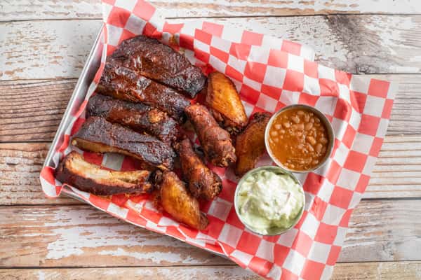 A platter of BBQ pork ribs and smoked chicken wings with coleslaw and a dipping sauce, served on red and white checkered paper