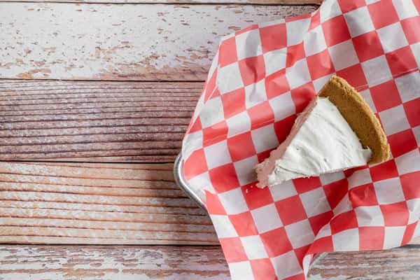 A slice of peanut butter pie with a whipped cream topping, served on red and white checkered paper on a rustic wooden surface