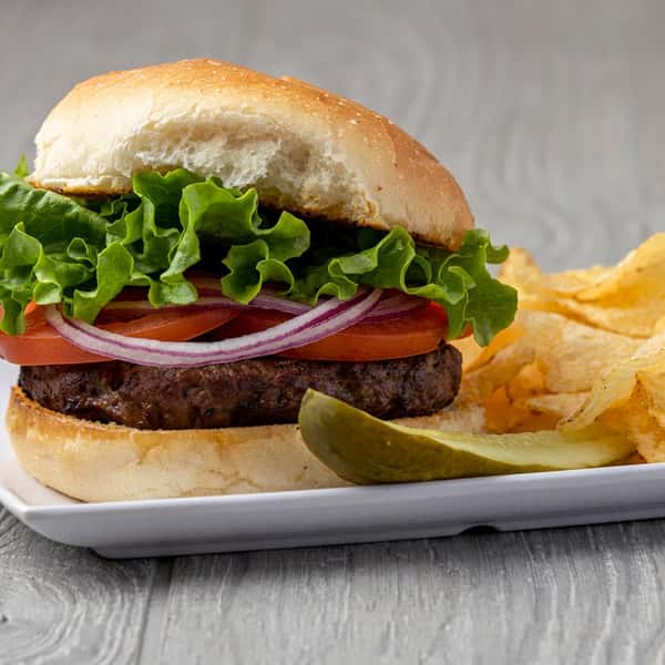 Hamburger with lettuce, tomato, and red onion, served with potato chips and a pickle spear on a white plate.